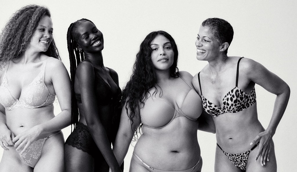 Four women of varying body types and skin tones stand together, smiling and embracing, wearing lingerie in neutral and leopard-print designs. The image emphasizes body positivity, diversity, and natural beauty with each woman displaying confidence and joy.