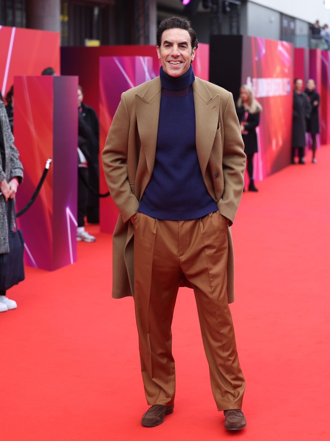 Sacha Baron Cohen in a brown suit and blue turtleneck jumper