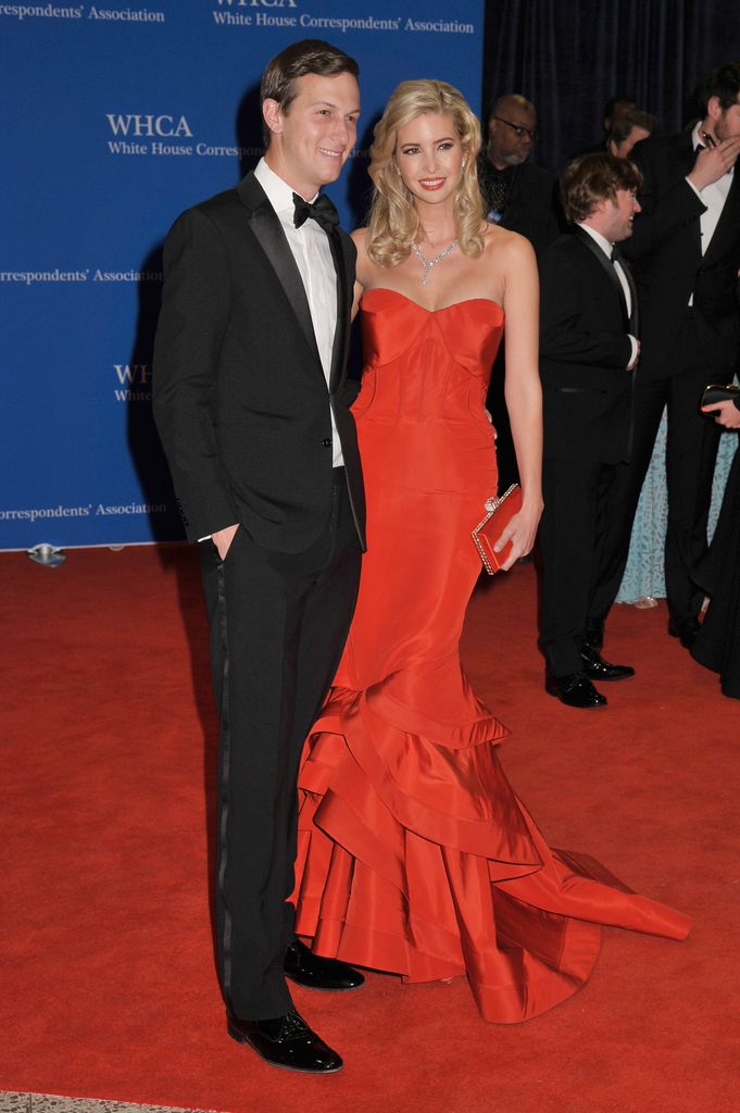 Ivanka wears a strapless red gown with a fitted bodice and layered skirt, paired with a diamond necklace. Her blonde hair is styled in soft waves, standing beside her husband in a tuxedo at the 101st Annual White House Correspondents' Association Dinner