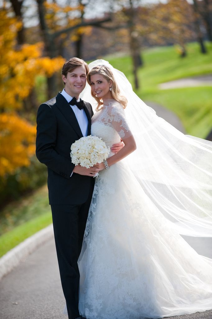 Ivanka wears a lace wedding gown with a long veil and white flower bouquet. She stands with her husband, both smiling, set against a sunny, leafy outdoor background.