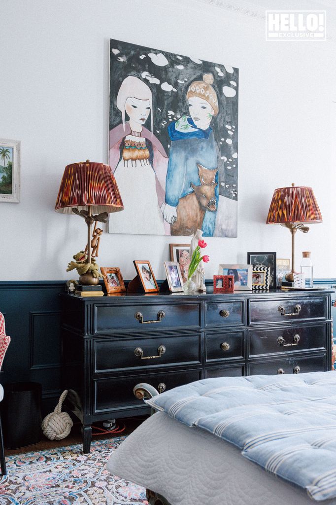 Nina Litchfield's bedroom at Notting Hill home. A bedroom with a dark blue dresser topped with two matching lamps and a large painting of two children. Family photos and a vase of tulips are also displayed on the dresser.