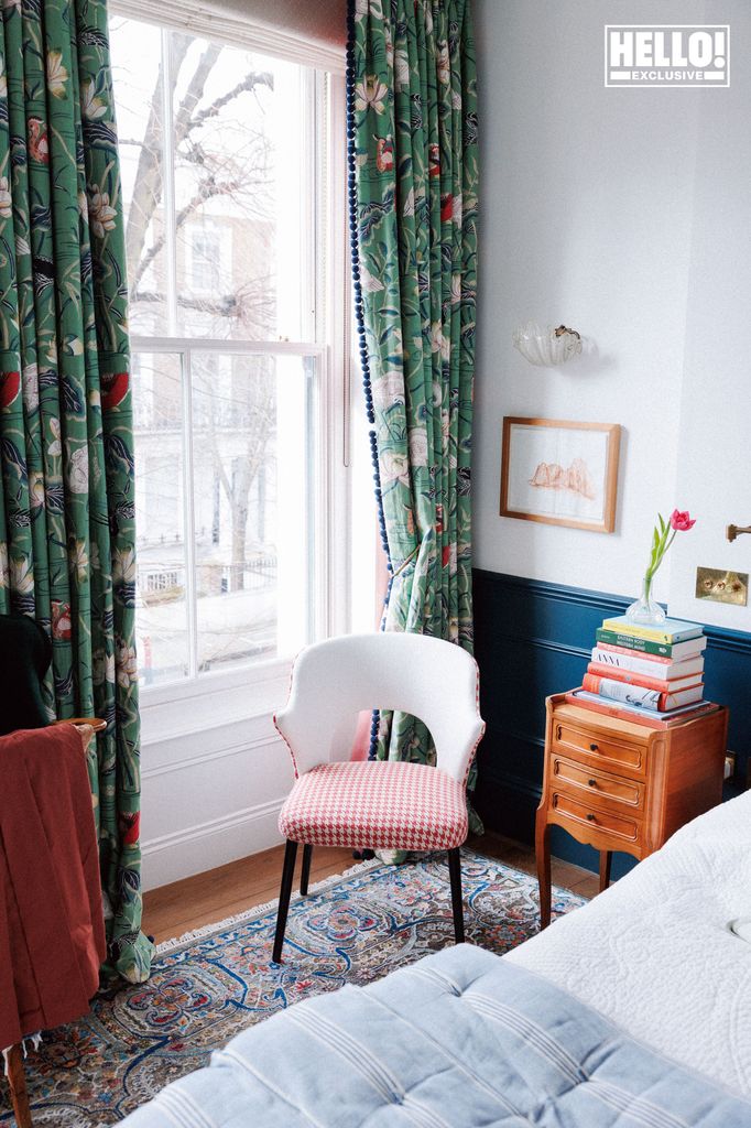 Nina Litchfield's bedroom at Notting Hill home. A bright bedroom with a large window framed by green curtains with a floral pattern. A gingham chair sits beside the window, and a wooden bedside table with books and a vase of tulips is next to the bed.