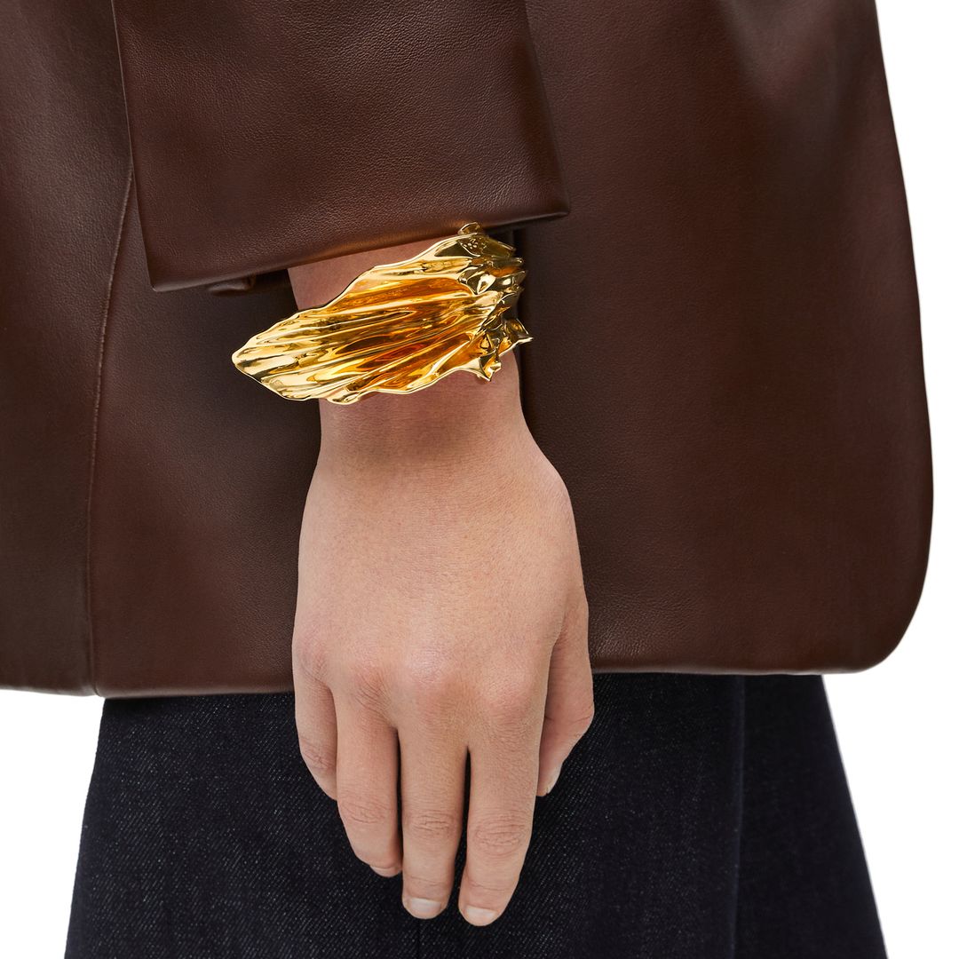 A person wearing a structured brown leather jacket with a striking sculptural gold bracelet peeking out from under the sleeve. The bracelet has an irregular, molten-like texture, creating a bold and artistic statement.