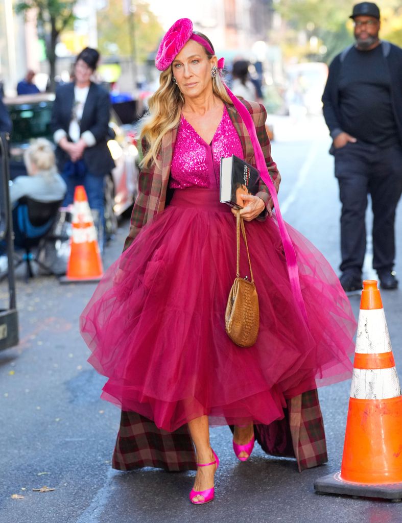 Carrie Bradshaw wears a vibrant pink sequined top with a voluminous magenta tulle skirt. She layers a plaid, oversized coat for contrast. Accessories include a bright pink fascinator, oversized earrings, a tan woven purse, and hot pink strappy heels. She holds a book, exuding confidence and signature bold style.