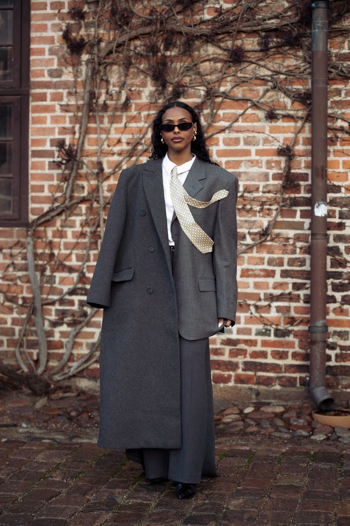 A guest wears grey pants, white shirt, grey jacket, yellow and green pattern tie and grey long coat outside the OpÃ©raSport show during the Copenhagen Fashion Week Autumn/Winter 2025 on January 27, 2025 in Copenhagen, Denmark