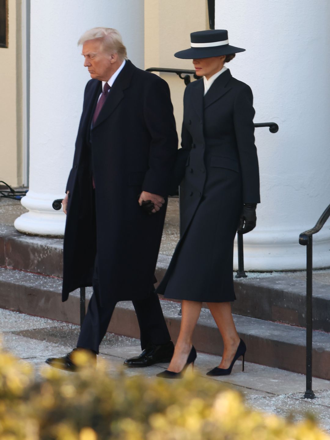 Melania Trump dressed in a sleek navy coat Paired with a dramatic wide-brimmed hat and pointy stilettos walking down the stairs next to husband Donald Trump