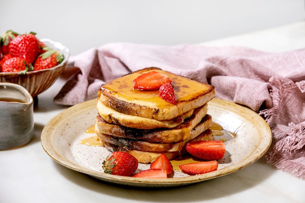 Stockpile of french toasts with fresh strawberries on ceramic plate. maple syrup in ceramic jug and pink cloth napkin over white table. Home breakfast