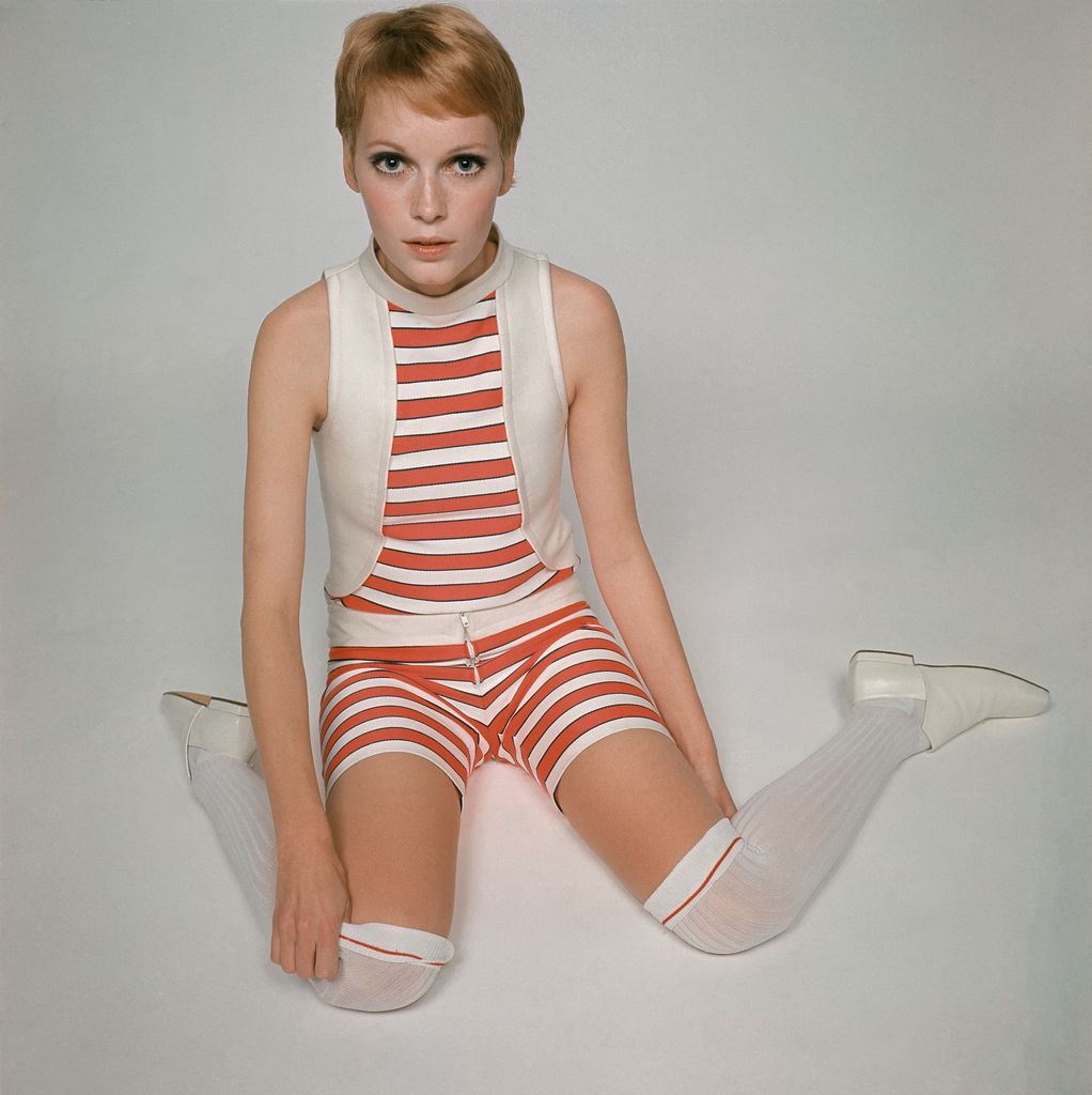 Mia Farrow kneeling on floor wearing a stretchy, striped top with matching, bicycle shorts and white knee socks, Glamour, October 1968