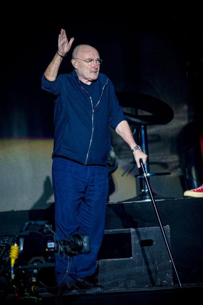 phil collins on stage holding a walking cain with one hand in the air