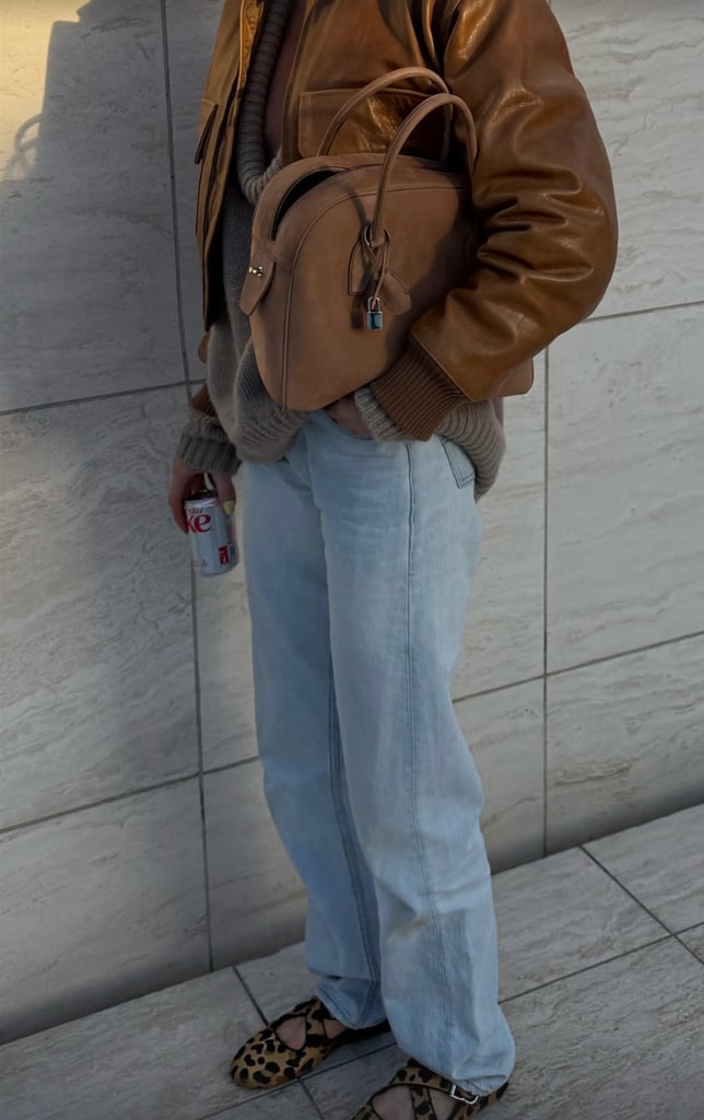 This image offers a closer look at the accessories and textures. The brown leather of Elsa Hosk’s jacket gleams under the light, showcasing its luxurious finish. The taupe handbag, held against her body, reveals intricate details—a subtle lock and fine stitching that signal high craftsmanship. Her Diet Coke can rests in one hand, a casual contrast to the refined elegance of her ensemble. The image captures the interplay of rich textures and muted tones, with the leopard-print flats peeking out from the soft, relaxed denim.