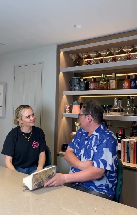 Alan Carr and Joanne McNally sitting at his kitchen island with bar shelf in the background behind them