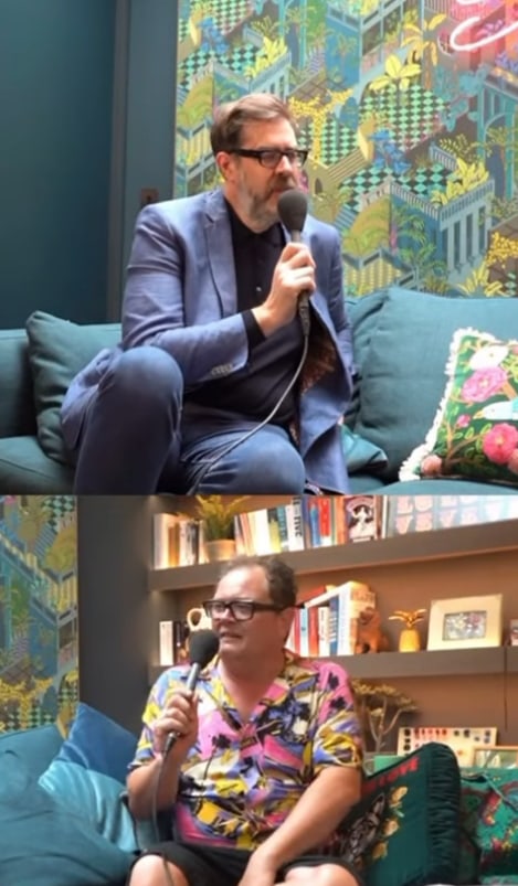 Alan Carr holding a microphone sitting on his sofa in front of shelves in his living room