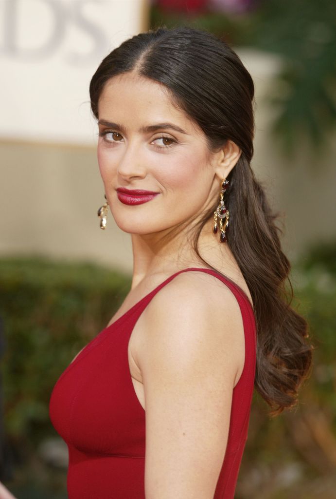 Salma Hayek in a red gown, wavy hair, red lipstick, and gold earrings radiating confidence and glamour.