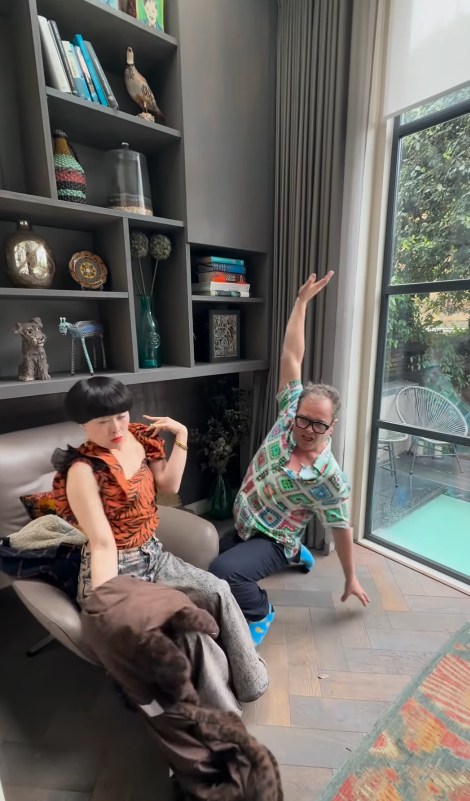 Alan Carr and Atsuko Okatsuka posing in his lounge with book case and window in the background