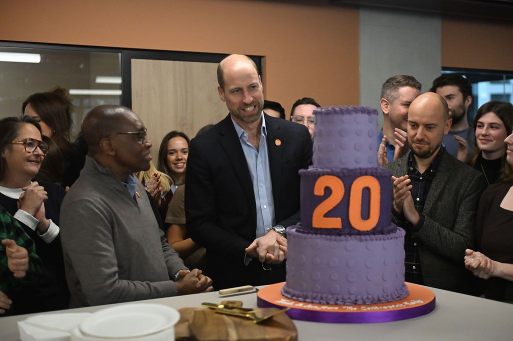 Prince William looking at a three-tier cake while people applaud