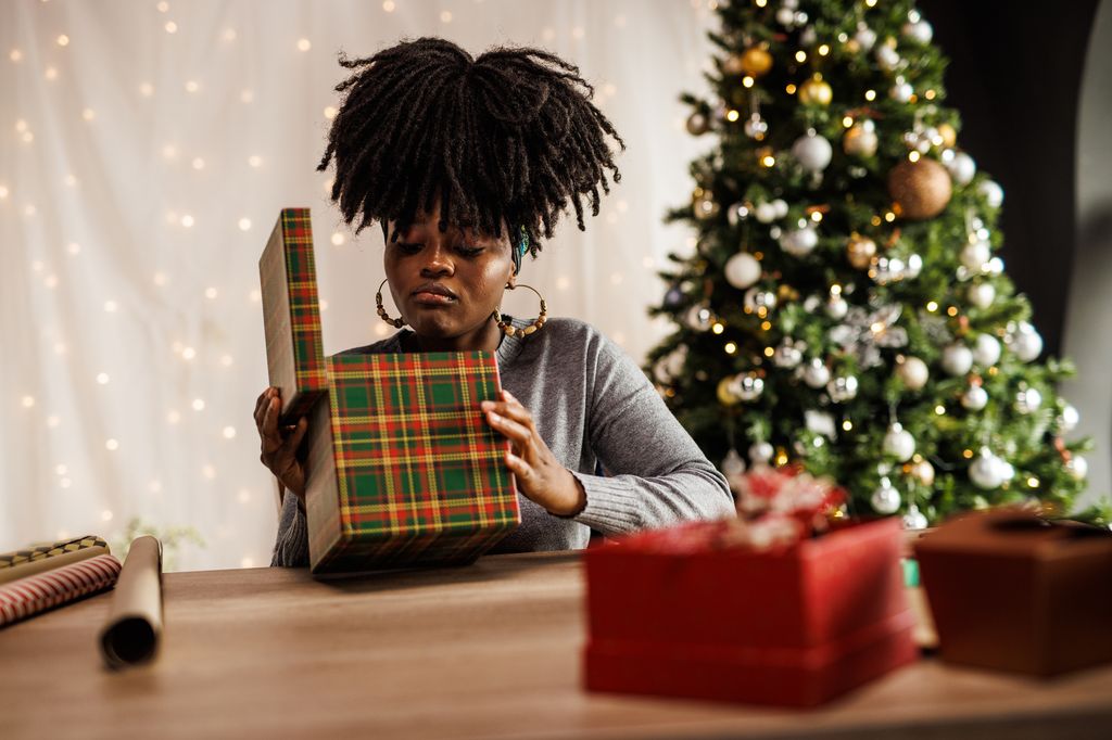 Portrait of disappointed young woman sitting at her home office desk and opening a Christmas gift box.