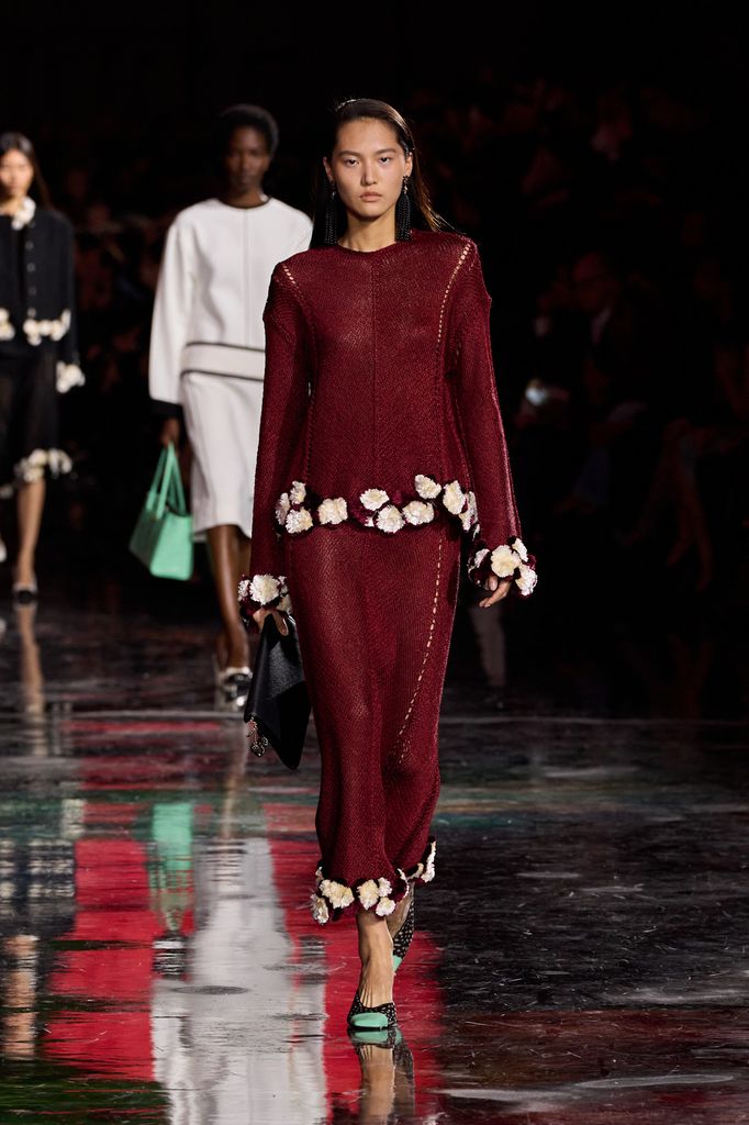 A model walks down the runway in a knitted top and matching skirt in burgundy with white floral detail on the hems