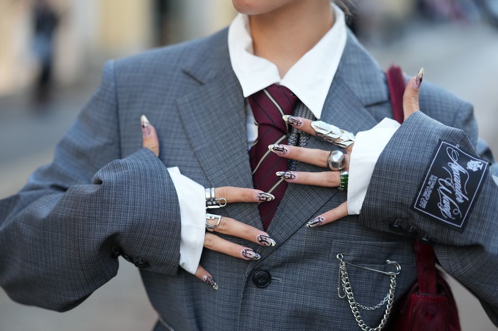 MILAN, ITALY - SEPTEMBER 25: A guest wears large multiple silver rings, a burgundy fuzzy shoulder bag, a white button-down shirt with visible collar, a burgundy and navy striped tie, a charcoal grey checked wool suit jacket with notch lapels, front flap pockets, a black sleeve patch and a silver chain draped from the pocket and worn as a belt, outside MM6, during Milan Fashion Week - Womenswear Spring/Summer 2026, on September 25, 2025 in Milan, Italy (Photo by Edward Berthelot/Getty Images)