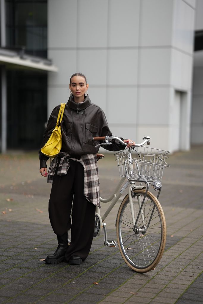 Evi Wave is seen wearing a dark brown oversized faux leather Aemmi bomber jacket with a high funnel collar, dropped shoulders, and exaggerated chest flap pockets; a longline brown and beige plaid flannel H&M shirt, tied asymmetrically around the waist; wide-leg, high-waisted dark brown flowing tailored COS trousers; she carries a yellow Balenciaga Le City bag maxi in Arena Storico lambskin with two leather hand-braided handles, brass hardware, front zipped pocket and knotted leather puller; on her fingers she wears silver rings from Ariane Ernst and her nails are polished in red