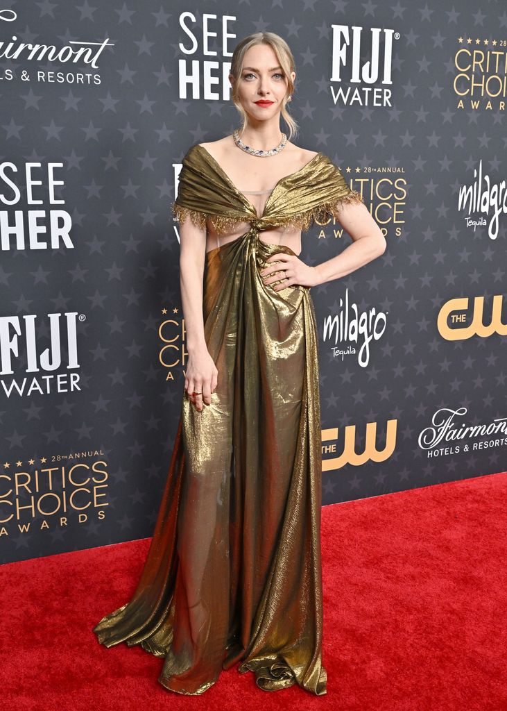 Amanda Seyfried wears a gold cut out gown with ruched shoulders. She poses with her hand on her hip, her blonde hair in a ponytail and bright red lipstick.
