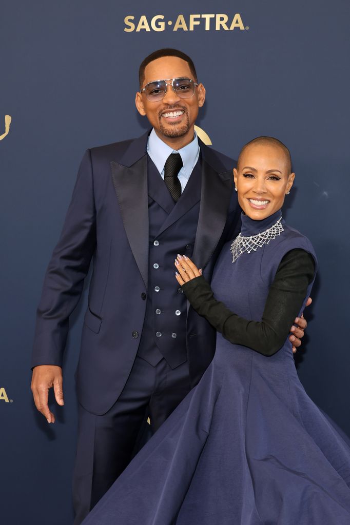 Will Smith wearing a suit and Jada Pinkett Smith wearing a long navy dress with black sleeves
