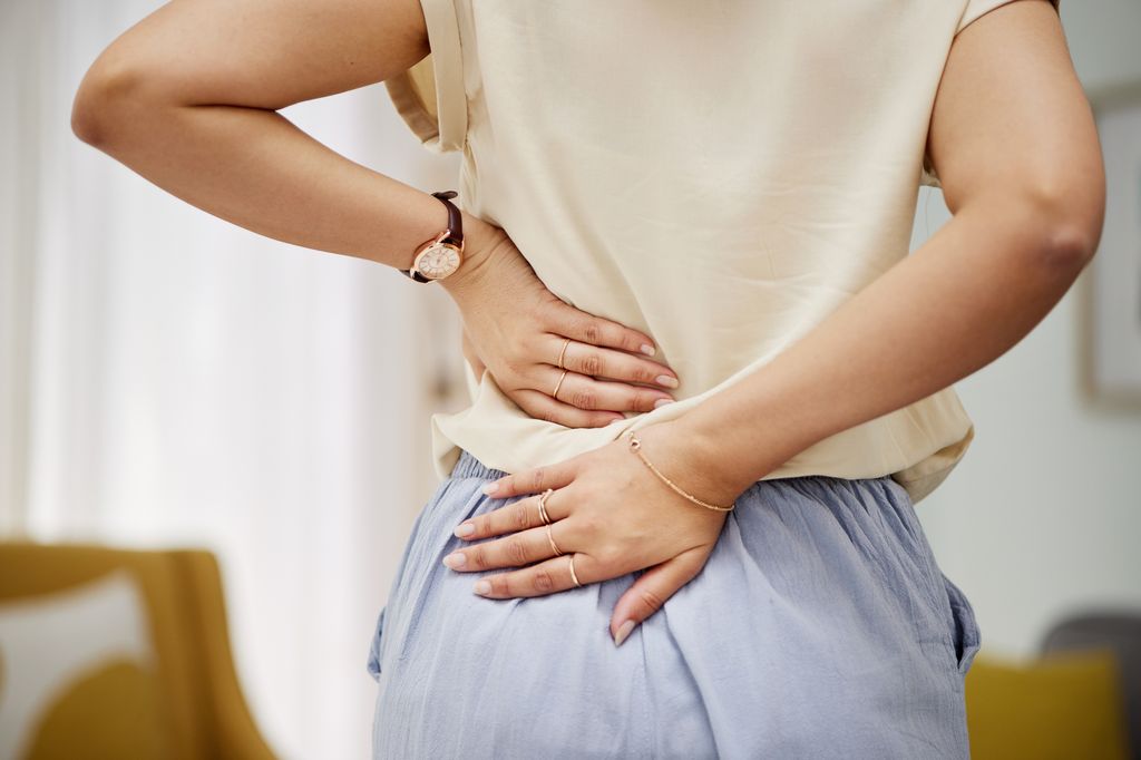 Close-up of a person in a beige shirt and light blue trousers pressing their hands against their lower back to indicate lumbar pain or discomfort.