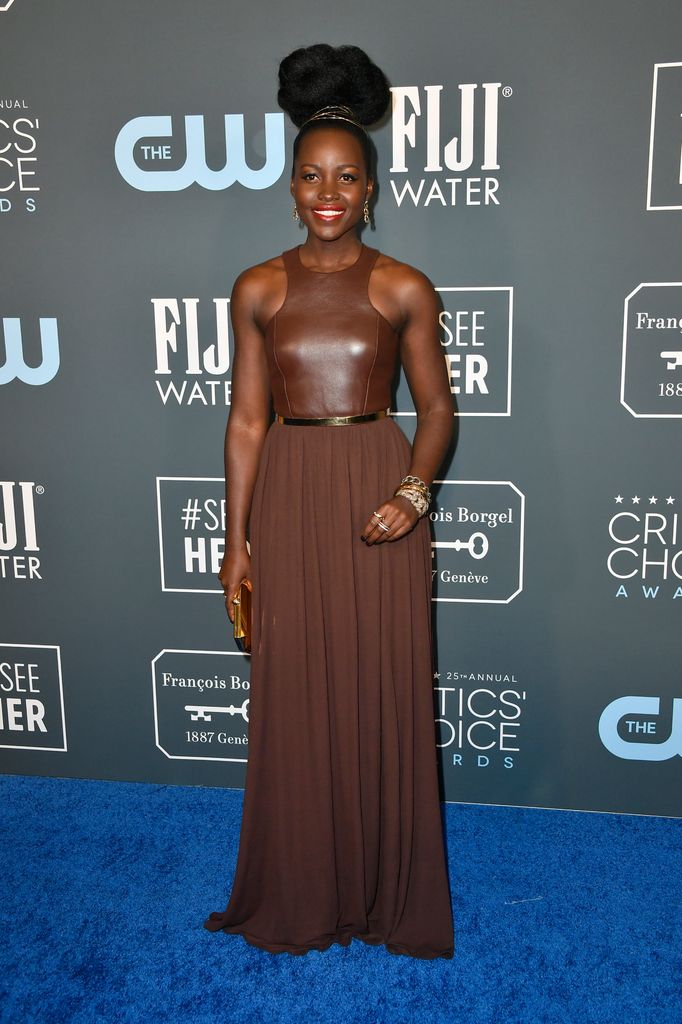 Lupita Nyong'o wears a brown halterneck dress with long flowing skirt and a thin gold belt