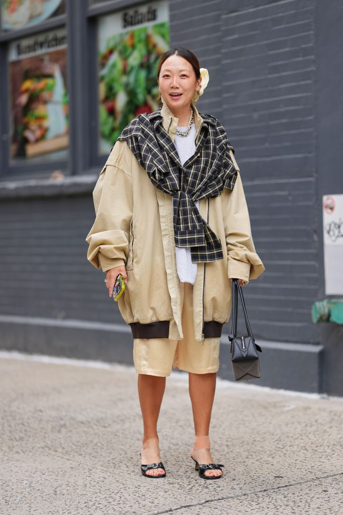A guest wears a center-parted low bun secured with a cream satin hair scrunchie, a chunky silver chain necklace, a black structured leather geometric top-handle bag, an oversized beige cotton coat with dark brown ribbed hem and cuff detailing, a black and beige plaid shirt draped and tied across the shoulders, a white crewneck T-shirt underneath, knee-length beige satin Bermuda shorts, and black patent leather open-toe mules with low block heels and front bow detailing