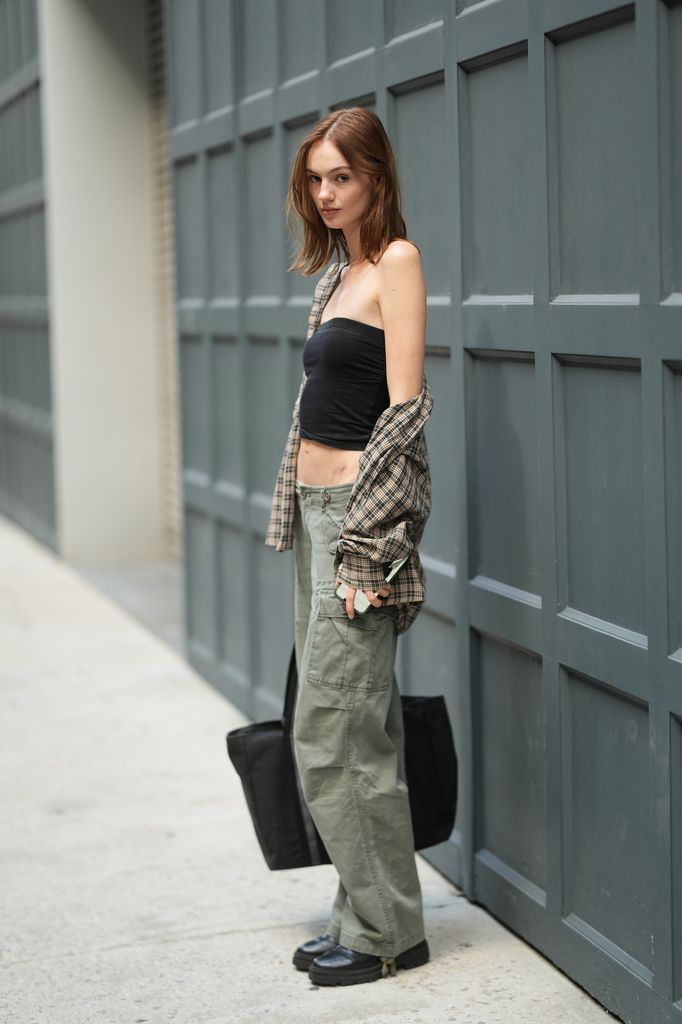 Reece Buchanan is seen wearing a black strapless top, plaid shirt over the shoulders, sage green cargo pants, black chunky shoes and carrying a black tote bag outside of the Ankvas show during New York Fashion Week