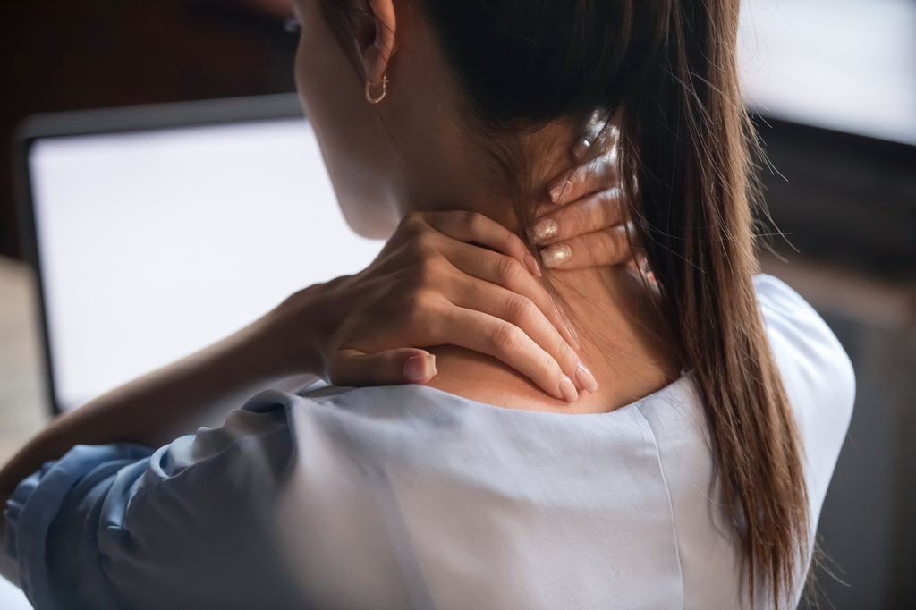 Close-up of a woman sitting in front of a laptop, holding her neck and shoulder with both hands to alleviate tension caused by computer use.