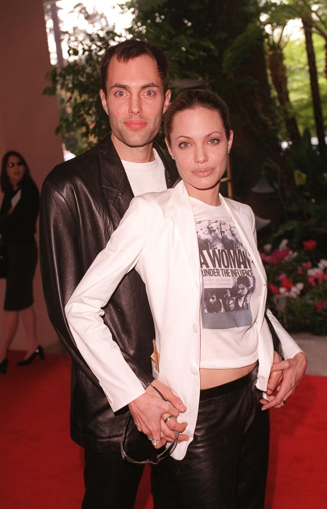 Angelina Jolie and her brother pose on the red carpet - he wears a black leather blazer, she wears a white blazer, leather trousers and a white and black tshirt. They hold hands. 