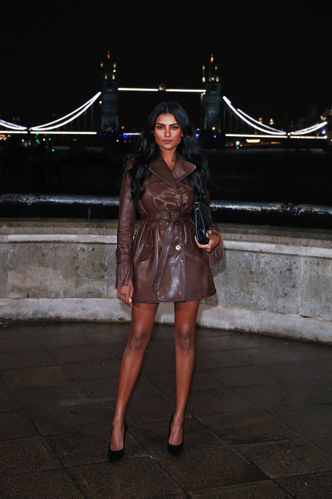 Simone Ashley wears a brown leather trench coat with black handbag and heels