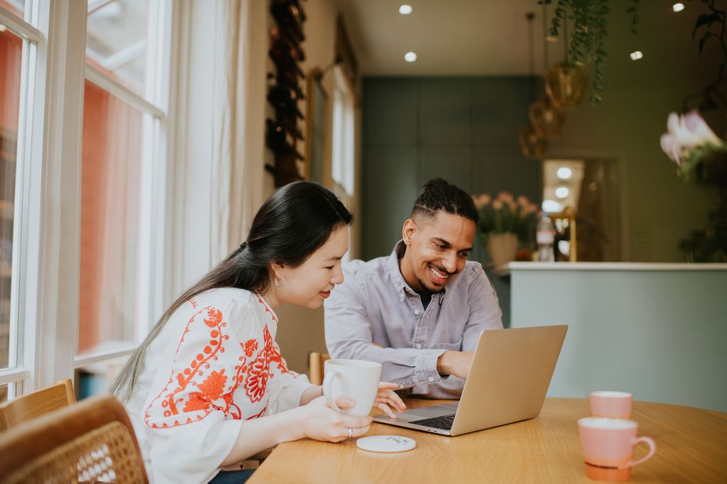 A black man and a Chinese woman sit at a table. They sit behind a laptop and look relaxed and have a lighthearted discussion. The man points at the computer screen.