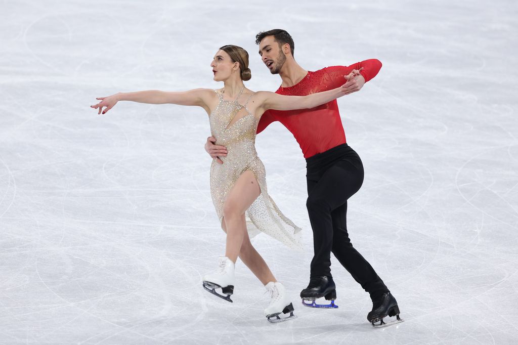  Gabriella Papadakis wearing a silver sparkly outfit and Guillaume Cizeron in a red top and black trousers, holding his partner around the waist on the ice  