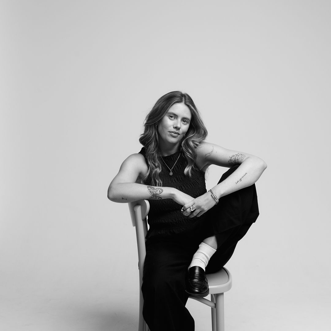 Black-and-white portrait of a woman with tattoos, sitting casually on a chair with one leg up, wearing a sleeveless top and wide-leg trousers.