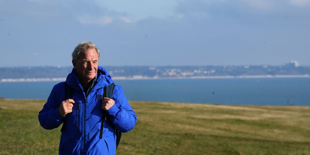 Man in blue rain coat and backpack standing on grass against sea backdrop