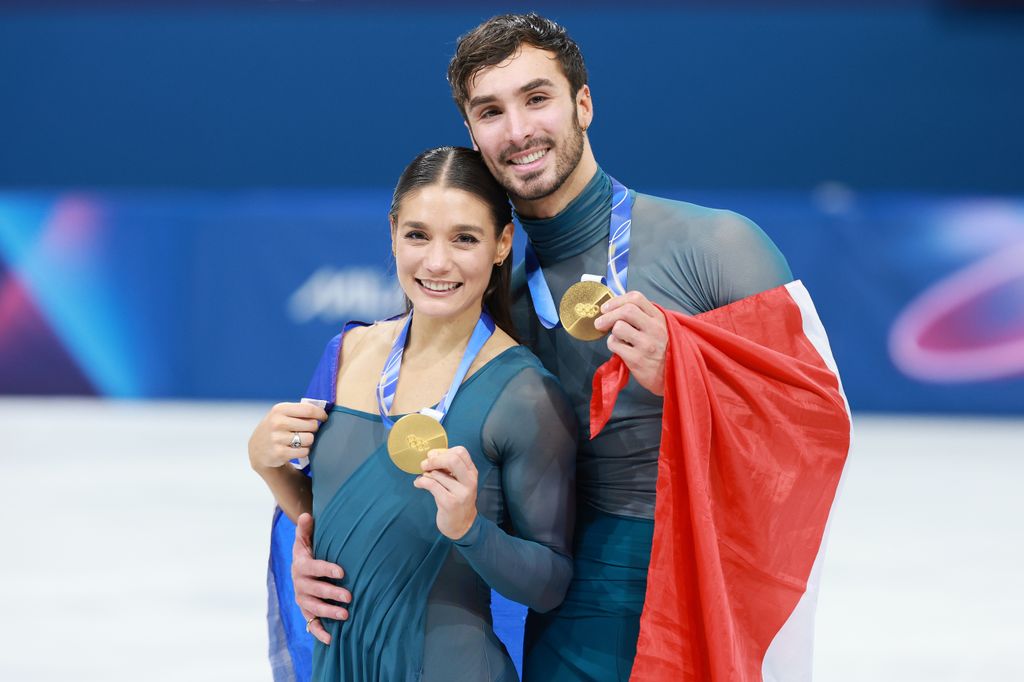 Laurence Fournier Beaudry and Guillaume Cizeron holding gold medals on the ice smiling, wearing the French flag and matching blue mesh ice-skating uniforms