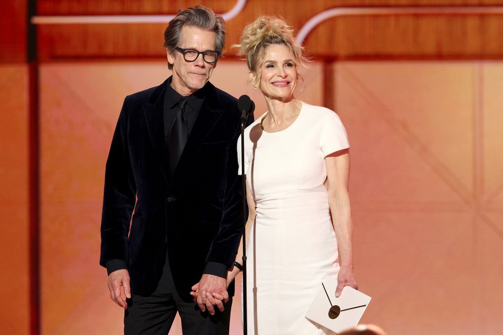 Kevin Bacon and Kyra Sedgwick speak onstage in black suit and white dress