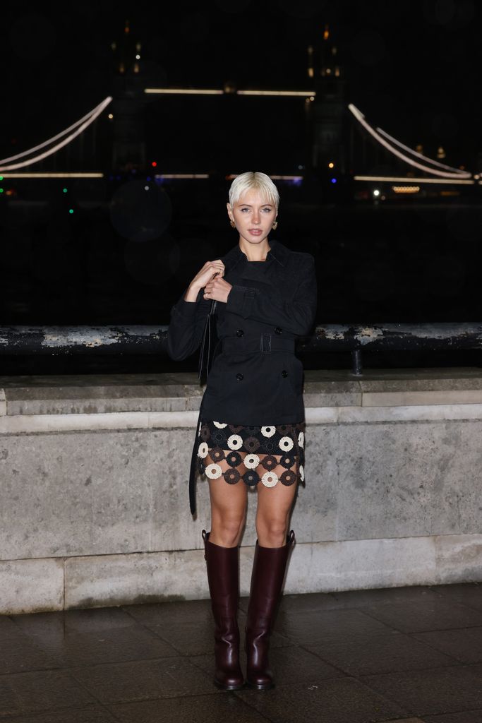 Iris Law wears a brown and cream crochet mini skirt, brown knee high boots and black trench coat