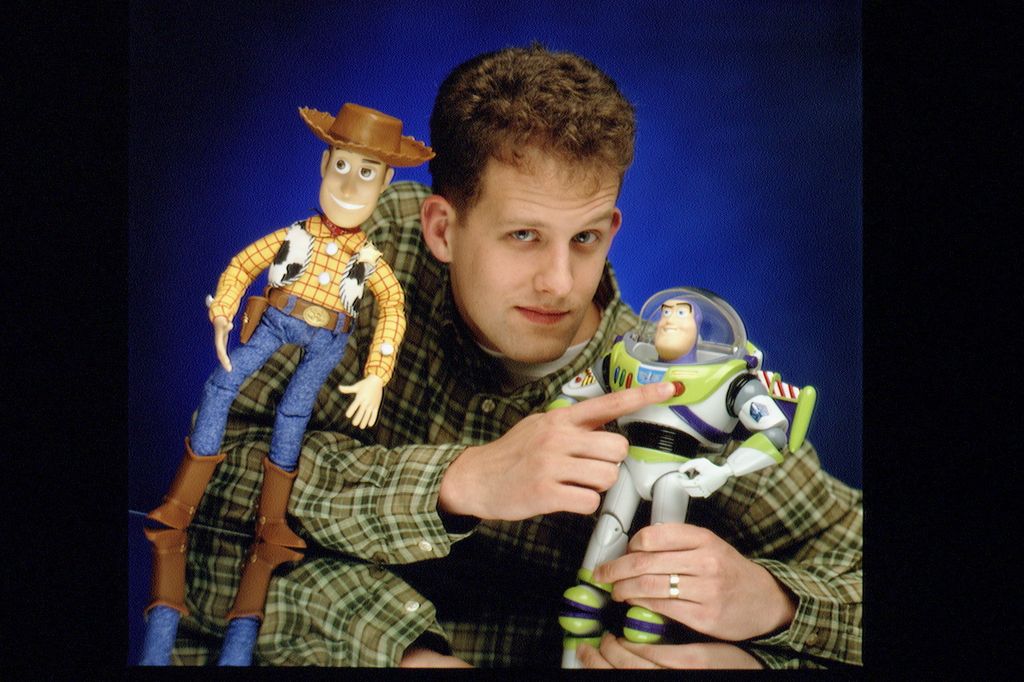 Man holds Buzz Lightyear toy in one hand and points to it with another while a Woody toy reclines against his arm.