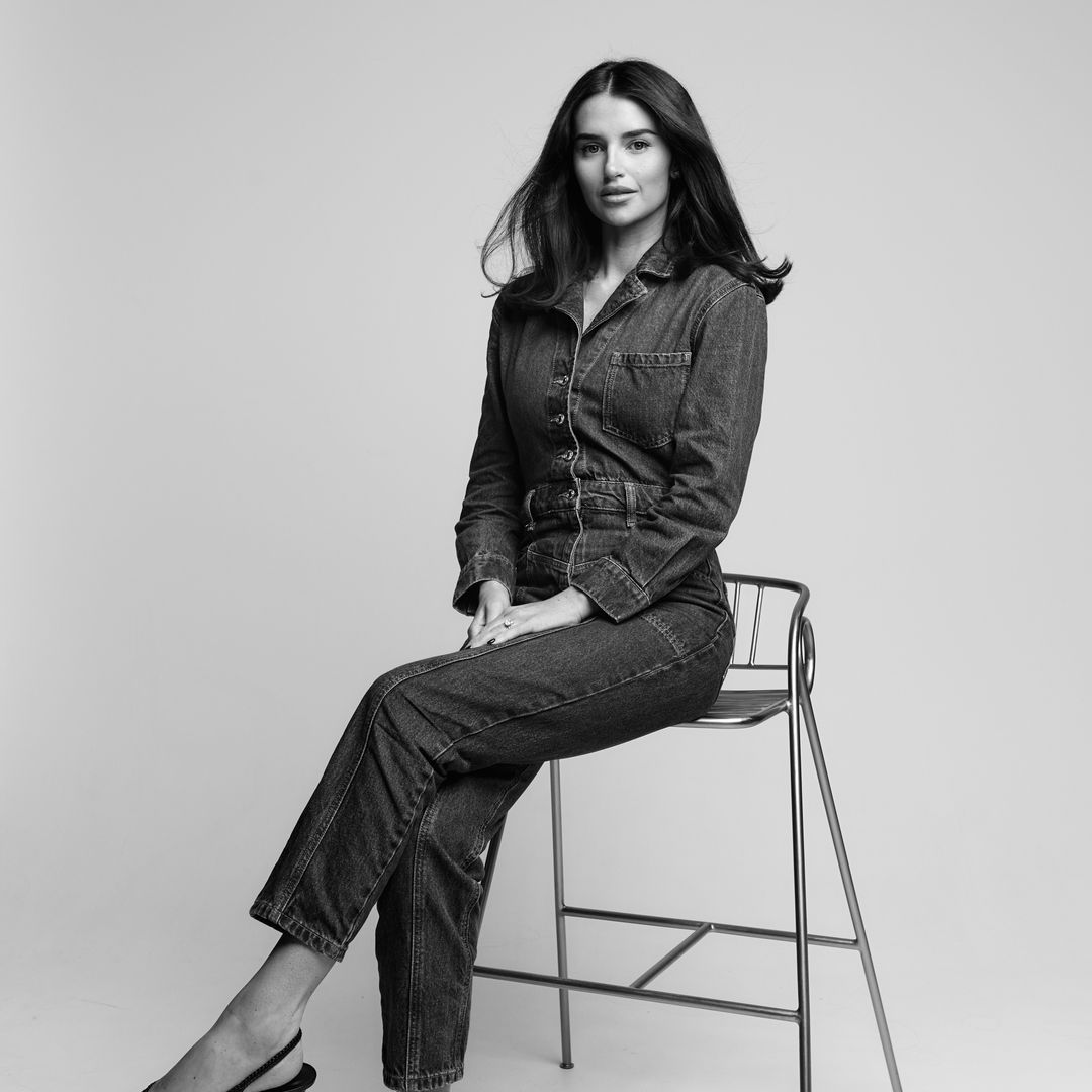 Black-and-white portrait of a woman wearing a denim jumpsuit and slingback heels, seated on a tall metal stool against a plain studio background.