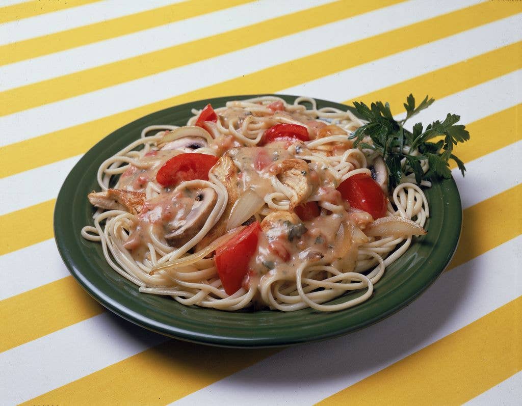 A dish of pasta noodles with tomatoes, chicken, onion and musroom pieces with a piece of parsley on the side, sits on a yellow and white striped surface.