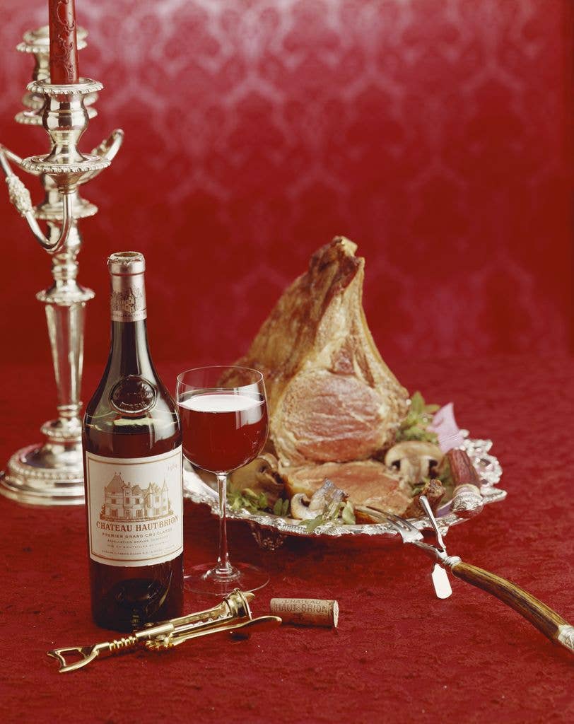 Red wine bottle, silver candelabra and a silver dish of cooked meat all resting on a red brocade background.