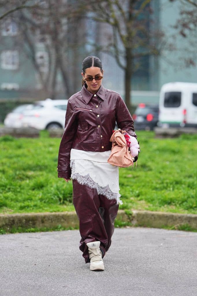 A guest wears black hair in a bun, brown tinted sunglasses, a dark burgundy shiny leather button front jacket, a white lace trimmed asymmetric top, matching dark burgundy shiny leather wide leg pants, a light beige leather nailed City handbag from Balenciaga, light beige wedge heels / ankle sneakers from Isabel Marant, outside Diesel, during Milan Fashion Week - Womenswear Fall/Winter 2026/2027, on February 24, 2026 in Milan, Italy (Photo by Edward Berthelot/Getty Images)