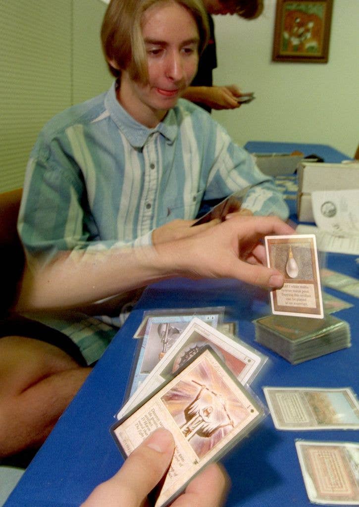 A man sits cross legged at a low blue table while an out of shot figure plays a Magic card onto the pile. 