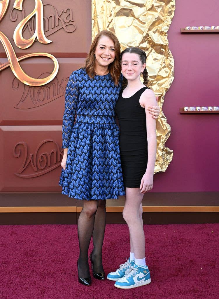 Alyson Hannigan in a knee length blue dress with black tights and black heels with her arm around daughter Keeva in a black all in one tank top and leotard with teal trainers.
