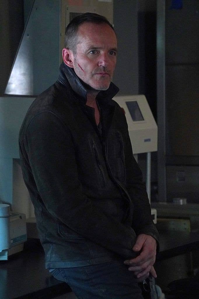 Clark Gregg as Agent Phil Coulson perching on a counter in jeans and a black zipped jacket with the collar popped up and a cut running across his jaw.