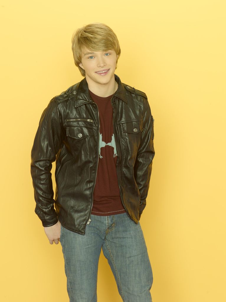 Sterling Knight stands with his hands hooked in his back pockets. He wears a dark red top, blue jeans and a black jacket. 