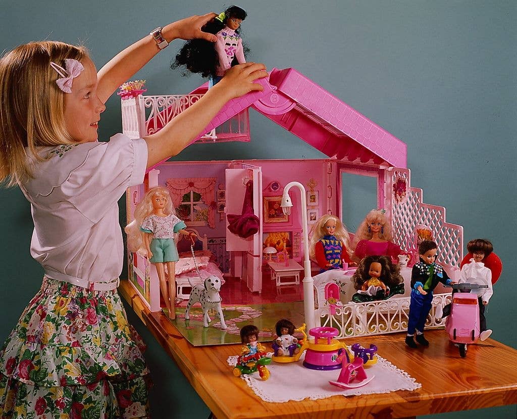 Girl with short blonde hair and a pink bow clip playing with a bright pink Barbie house set where various dolls are shown sitting on chairs or playing with friends.