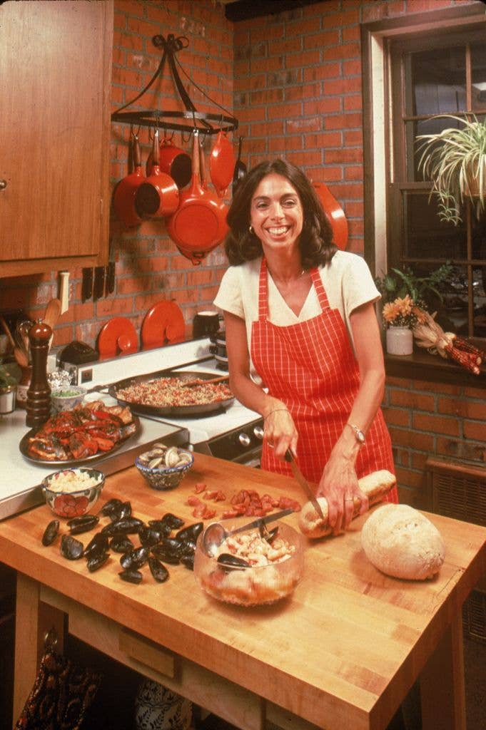 A smiling brunette woman cuts bread next to mussels on a chopping block in a kitchen as a large pan simmers on the stove.